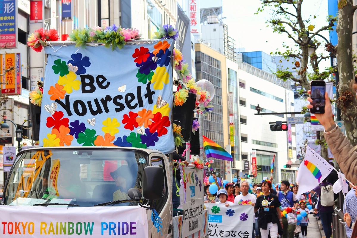 Tokyo Rainbow Pride parade float decorated with colorful flowers and a large “Be Yourself” sign, moving through a busy Tokyo street with marchers carrying rainbow flags behind it.
