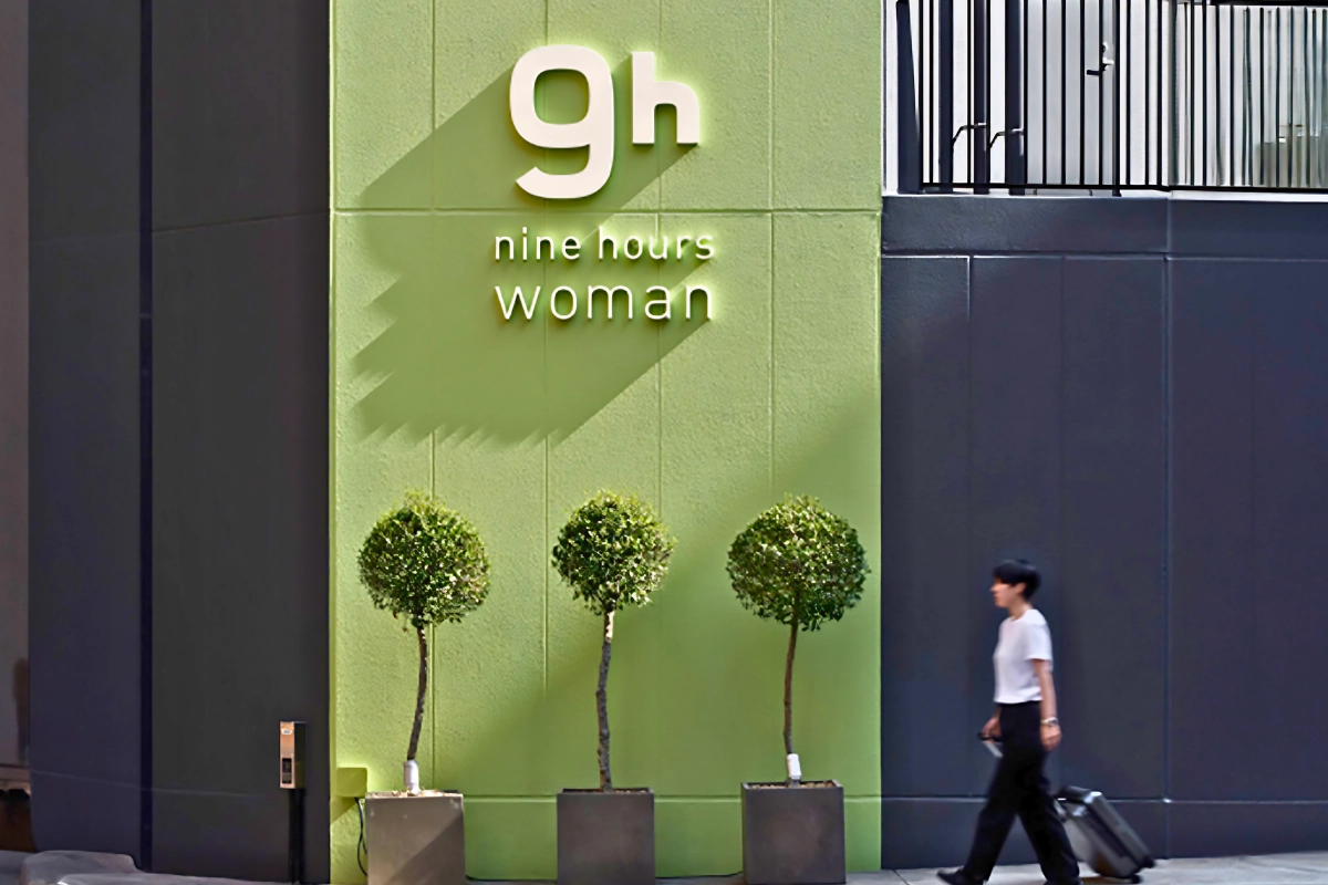 Best Capsule Hotels Near Tokyo’s Ni-chome for LGBTQ+ Travelers 14 Exterior of a Nine Hours women’s capsule hotel, featuring a green “9h nine hours woman” sign, three potted trees, and a traveler walking past with luggage.