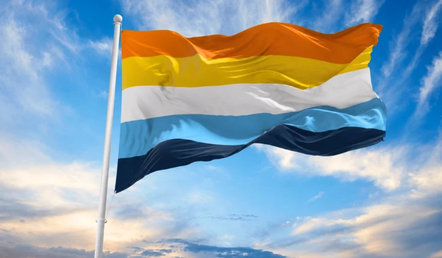 Sunny sky behind the sunset aroace flag waving in the wind attached to a white flag pole.
