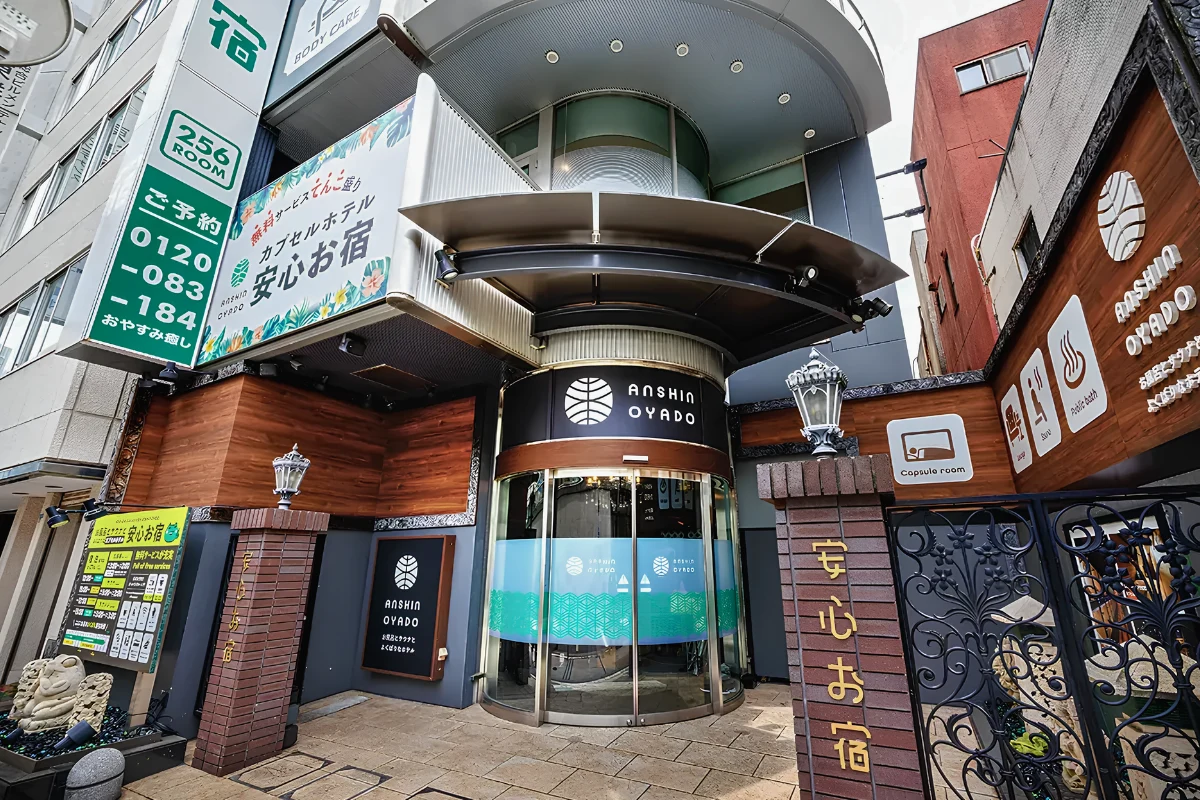 Best Capsule Hotels Near Tokyo’s Ni-chome for LGBTQ+ Travelers 13 Exterior of Anshin Oyado capsule hotel in Japan, showing the entrance, Japanese signage, and icons for capsule rooms, sauna, and public bath facilities.