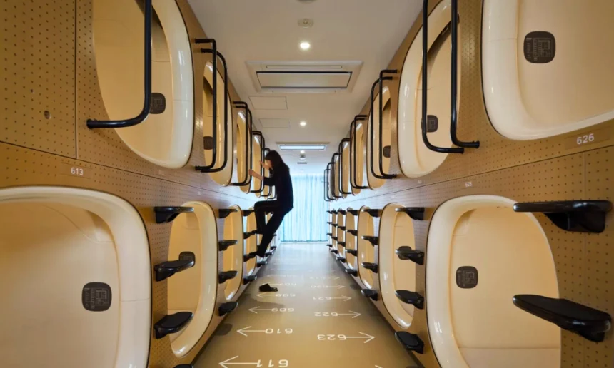 Rows of stacked sleeping capsules line a narrow corridor inside a Japanese capsule hotel, with a guest climbing into one of the pods.