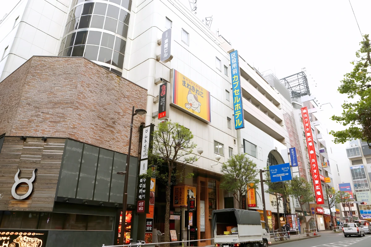 Best Capsule Hotels Near Tokyo’s Ni-chome for LGBTQ+ Travelers 12 Exterior of a capsule hotel building on a busy Japanese city street, with large vertical signs advertising the hotel among surrounding shops and businesses.