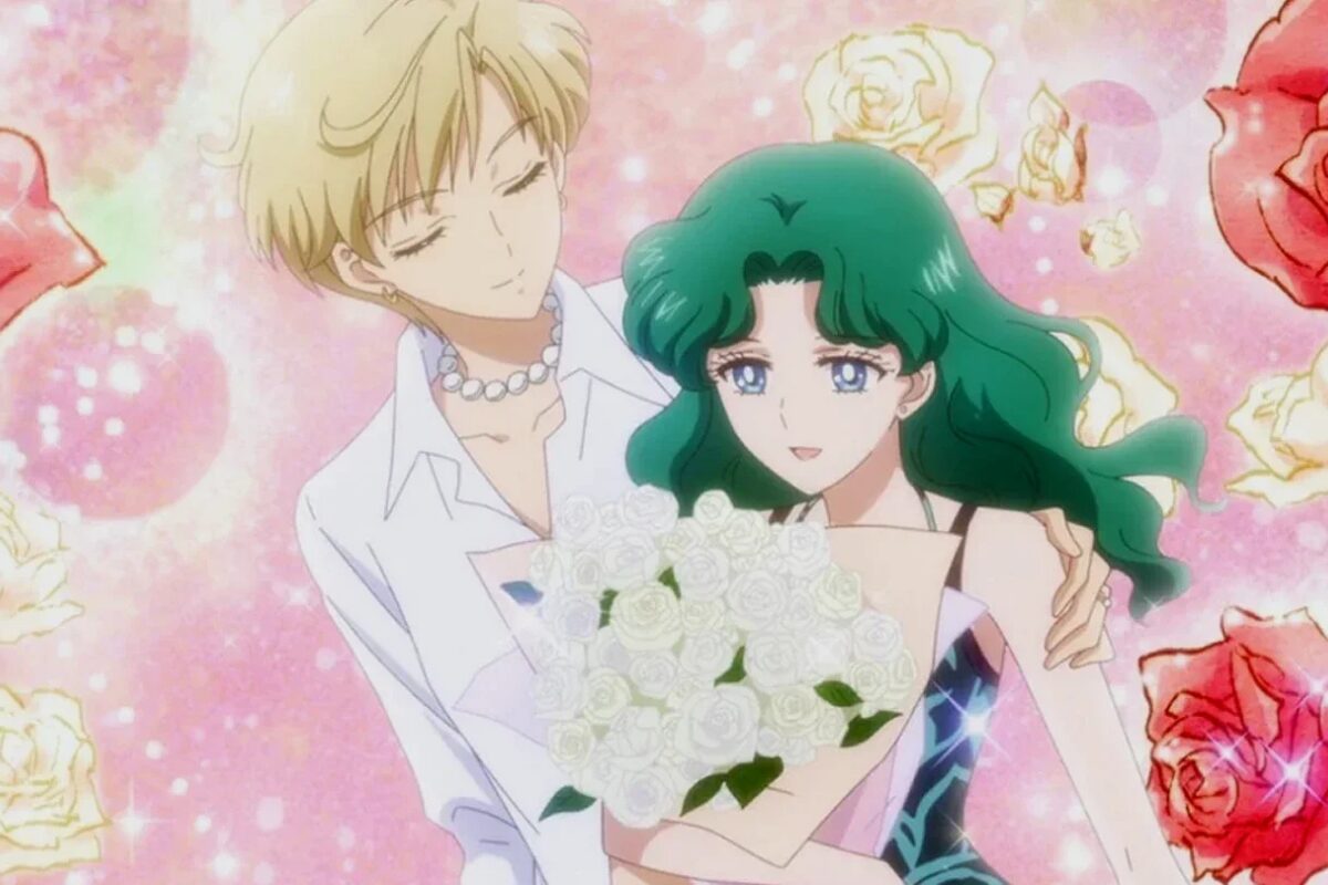 Haruka Tenoh, left, wearing a white shirt and pearls, puts her left arm around her partner, Michiru Kaioh, wearing a black dress and holiding a bunch of white flowers.