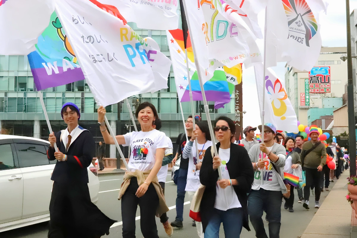 Pride parade participants are walking down the street waving large pride flags and smiling