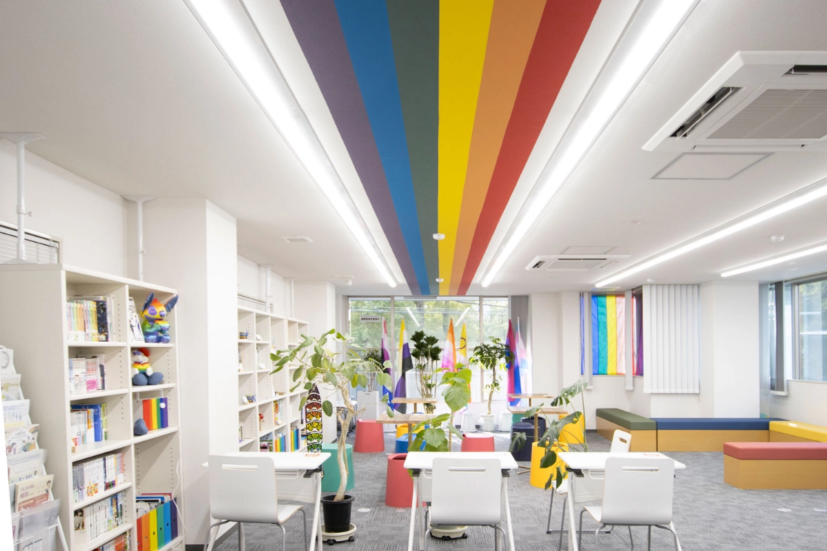What Is It Like to Be Bisexual in Japan? 18 Interior of a bright LGBTQ+ community space with rainbow stripes across the ceiling, Pride flags, bookshelves, plants, tables, and colorful seating.