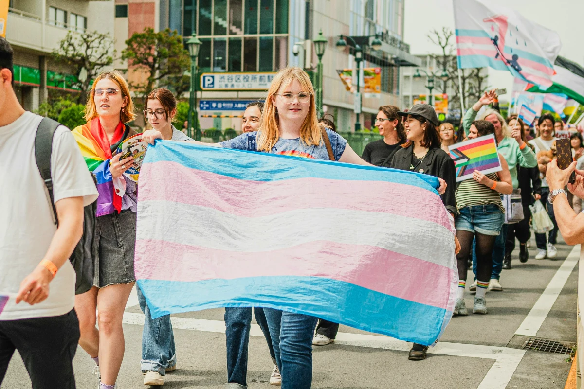 Pride parade is walking through the streets waving various pride flags. One person is seen holding open the trans pride flag and looking into the camera