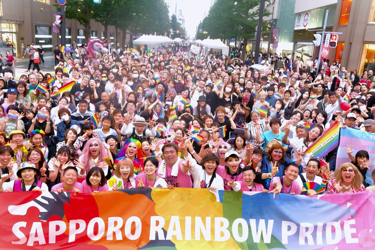 Participants of Sapporo Rainbow Pride pose for photo together. Large crowd is gathering round a banner waving pride flags 