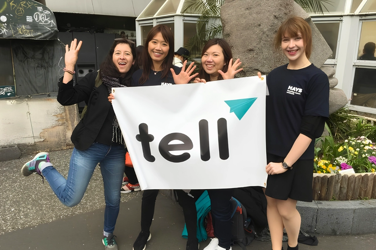 Four people smile and pose with a large TELL banner at an outdoor public event in Japan.
