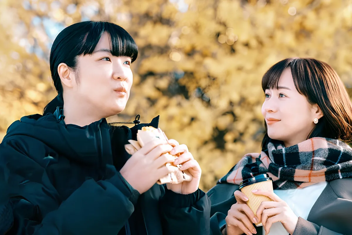 10 Best Japanese Lesbian Movies & Series to Watch 18 Two women sit together outside, one eating a sandwich and the other drinking a coffee.
