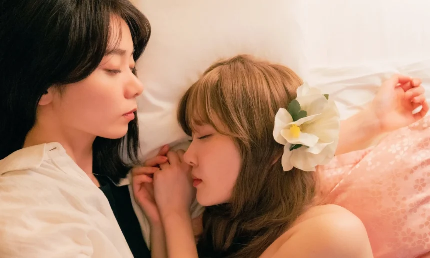 Two women from the Japanese lesbian tv show “Aya Is in Love with Hiroko!” lie closely together on a bed, one resting against the other with their hands clasped, while a large white flower decorates one woman’s hair.