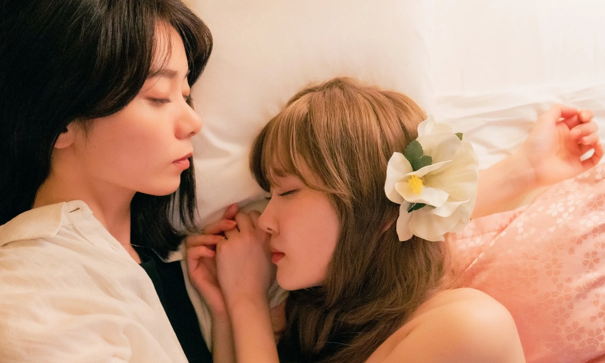 Two women from the Japanese lesbian tv show “Aya Is in Love with Hiroko!” lie closely together on a bed, one resting against the other with their hands clasped, while a large white flower decorates one woman’s hair.
