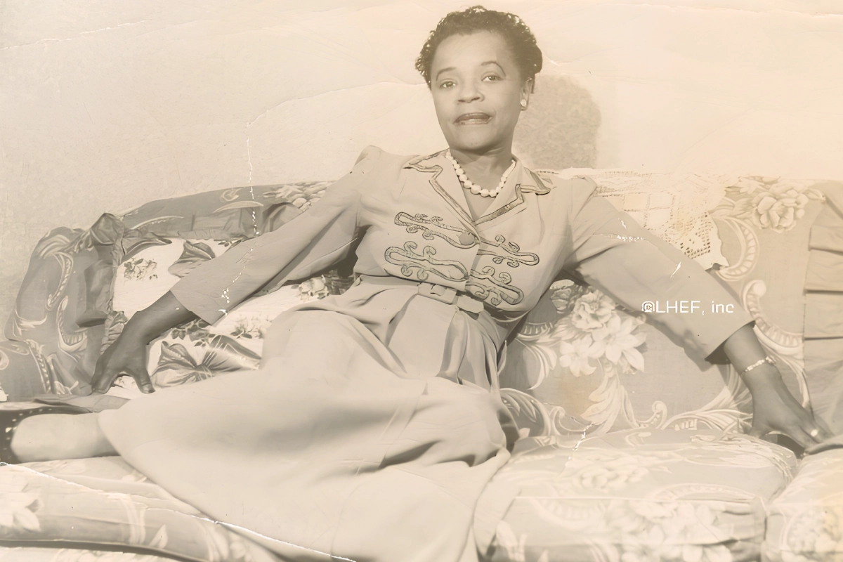 Vintage black-and-white portrait of a Black woman reclining on a floral sofa, wearing an embroidered dress and pearl necklace.