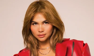Studio portrait of a feminine-presenting person with long light-brown hair, glossy makeup, layered necklaces, and a bright red leather jacket against a pale background.