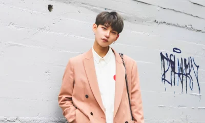 Androgynous-presenting young person with short dark hair and earrings, wearing a brown blazer and white shirt while looking directly at the camera.