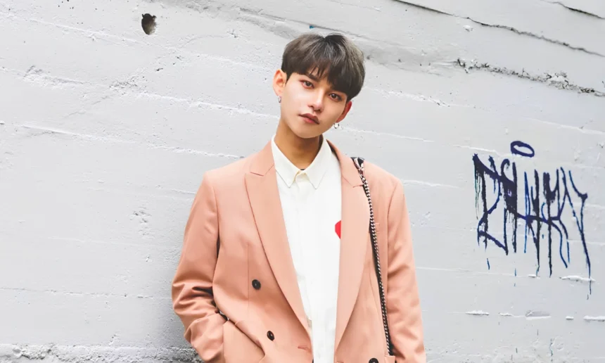 Androgynous-presenting young person with short dark hair and earrings, wearing a brown blazer and white shirt while looking directly at the camera.