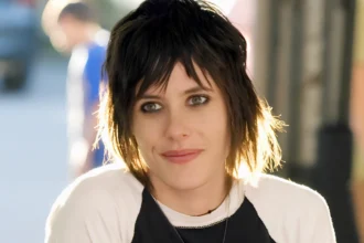 Close-up portrait of an androgynous-presenting person with short, layered dark hair and smoky eye makeup, wearing a black-and-white top in soft outdoor light.