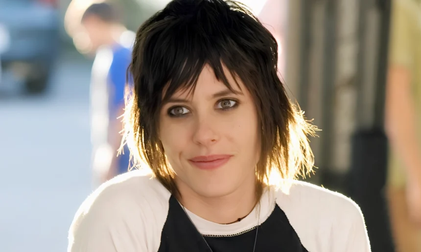 Close-up portrait of an androgynous-presenting person with short, layered dark hair and smoky eye makeup, wearing a black-and-white top in soft outdoor light.