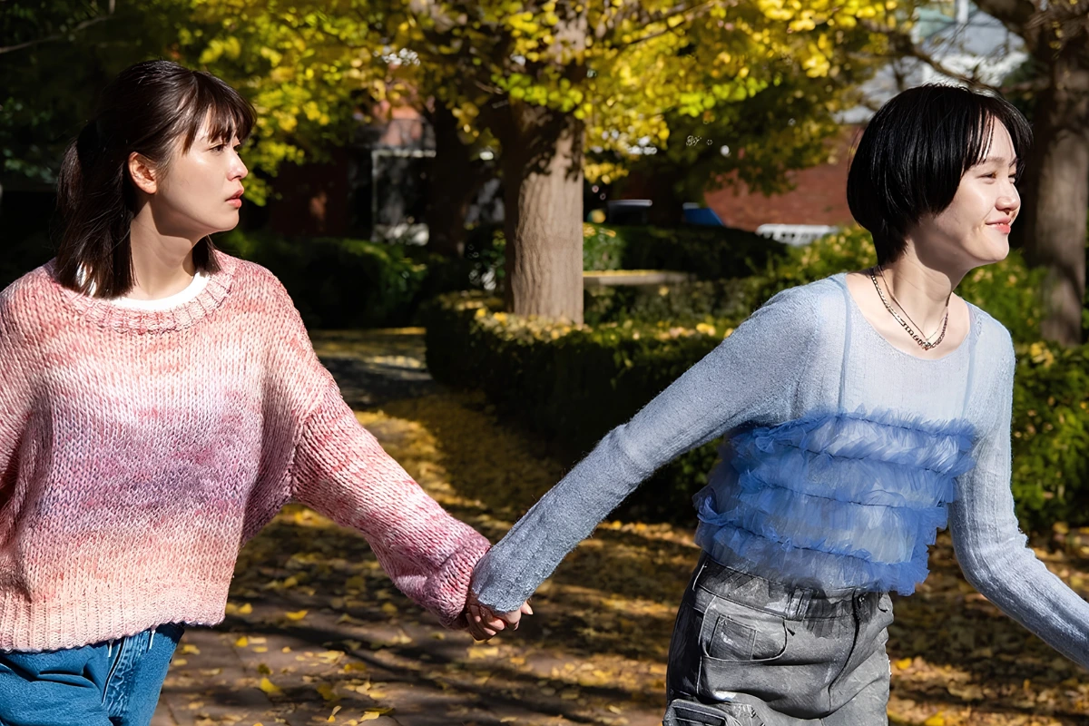 10 Best Japanese Lesbian Movies & Series to Watch 21 Two women hold hands while walking through a sunlit park, one in a pink sweater looking toward the other, who smiles while leading her forward.