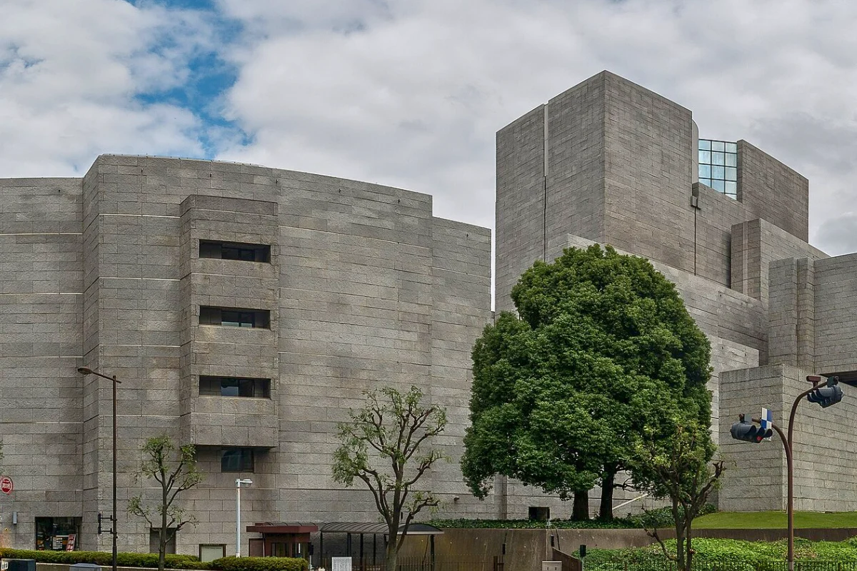 Exterior of the Supreme Court of Japan in Tokyo, showing its large geometric stone façade with trees in the foreground beneath a cloudy sky.