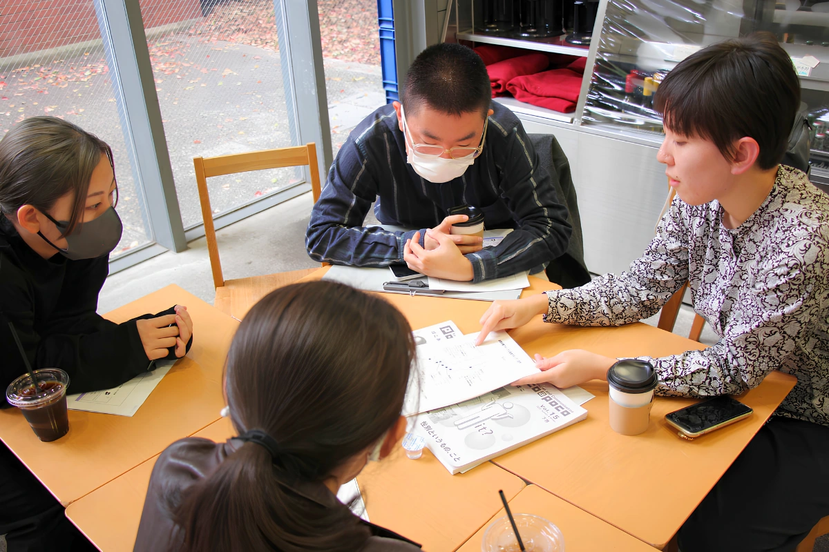 Four people sit around a table in a bright room discussing printed materials, with drinks, notebooks, and a smartphone on the table.