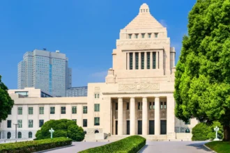 Exterior of Japan’s National Diet Building in Tokyo, framed by green trees and hedges beneath a clear blue sky.