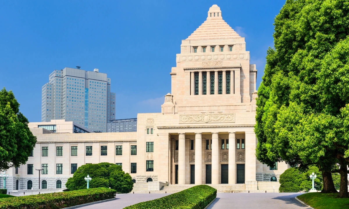 Exterior of Japan’s National Diet Building in Tokyo, framed by green trees and hedges beneath a clear blue sky.