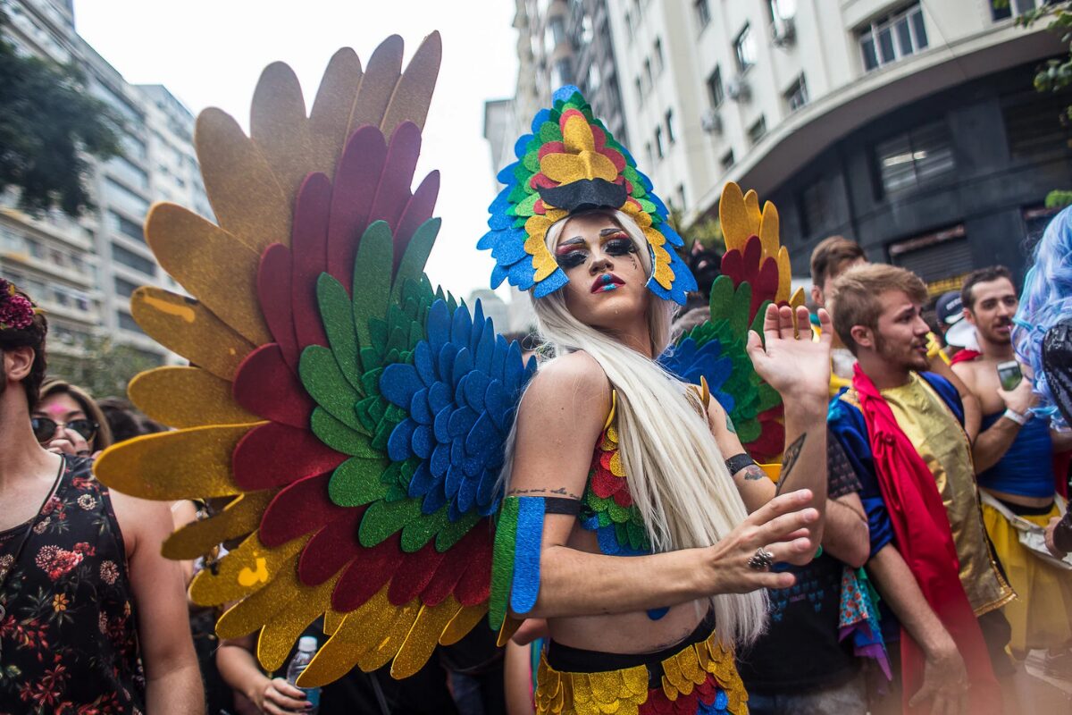 A drag queen in a rainbow-colored bird costume looks piercingly towards the camera