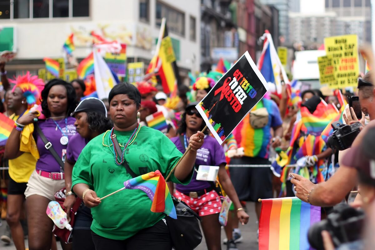 Marchers walk through Toronto, holding rainbow flags. At the front, one woman holds a sign reading “Get Tested to Feel Rested”