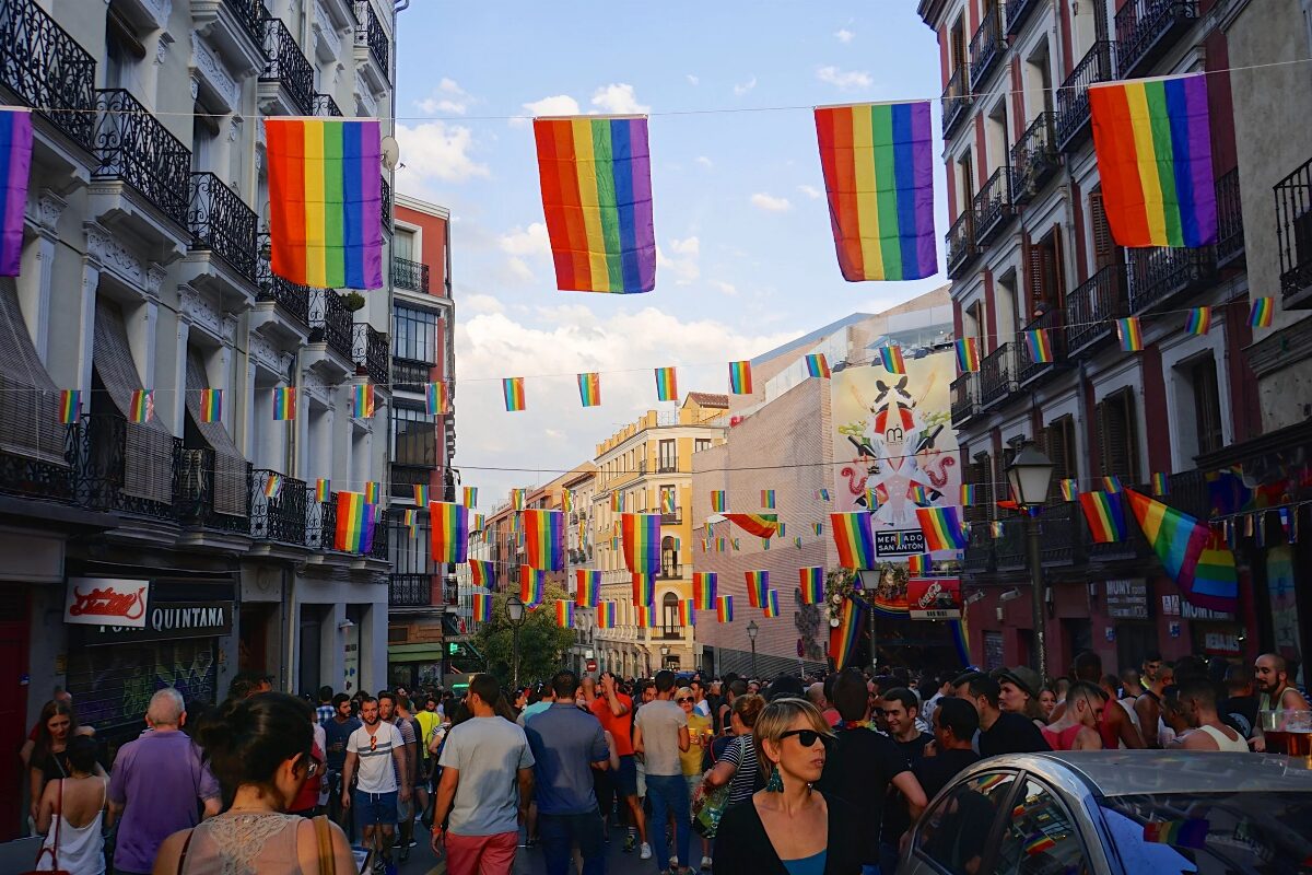 Marchers walk through a street in Madrid. Rainbow colored flag bunting is hung up stretching from one side of the street to the other