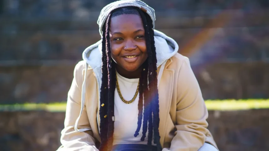 A smiling Black stud lesbian demonstrating stud fashion and style, wearing a tan coat, hoodie, beanie, and gold chain necklace.