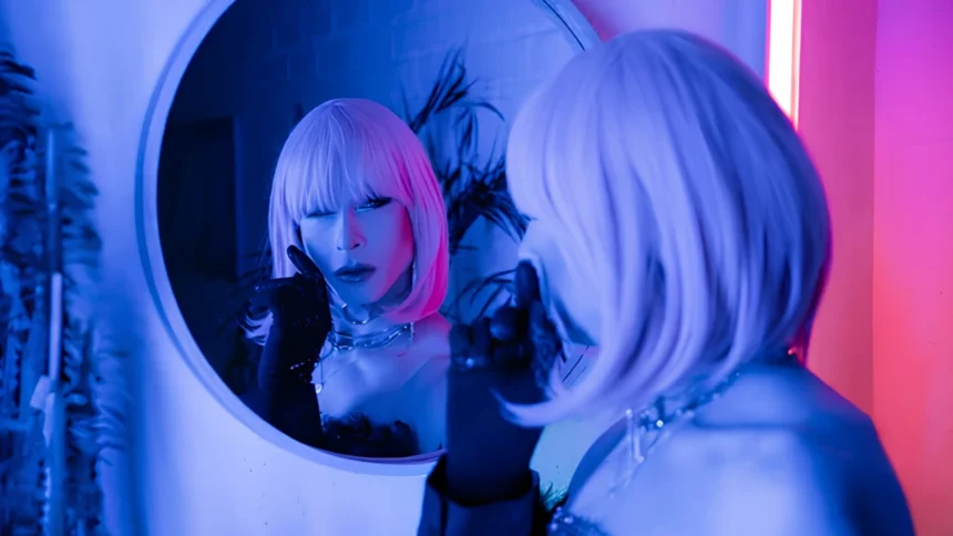 Performer in a platinum wig and dramatic makeup poses before a neon-lit mirror.