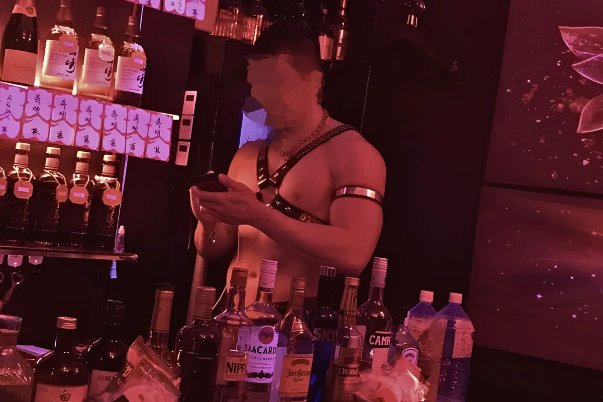 Muscular shirtless performer wearing a harness stands behind a bar under red nightclub lighting.