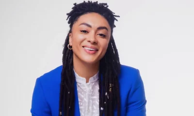 Smiling Black person with long locs wearing a bright blue blazer and white blouse, photographed against a plain light background.
