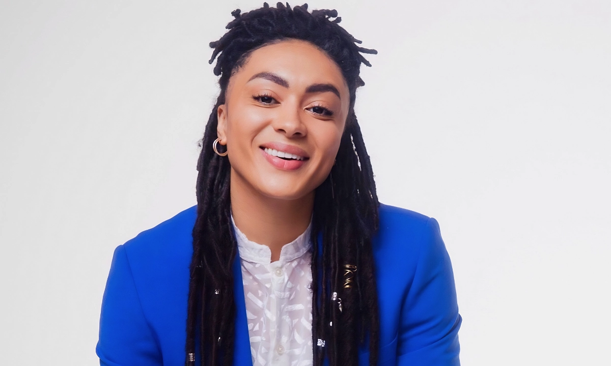 Smiling Black person with long locs wearing a bright blue blazer and white blouse, photographed against a plain light background.