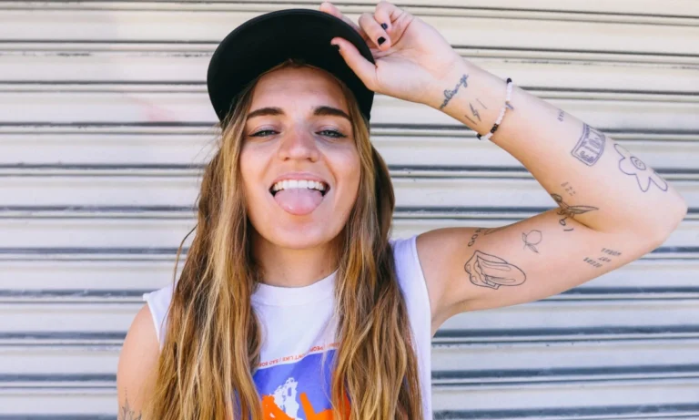 Smiling person with long hair and visible arm tattoos wears a black baseball cap and sleeveless top while posing in front of a metal shutter.