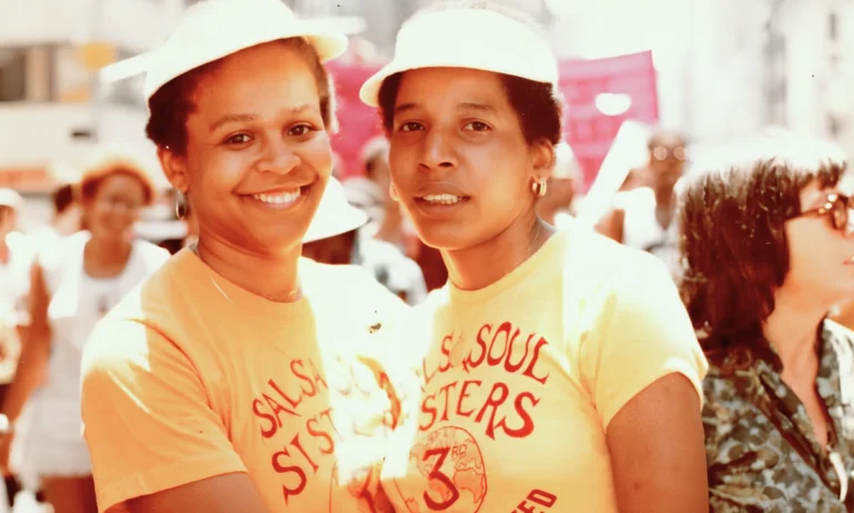 Two women wearing matching yellow t-shirts and hats stare into the camera and smile together in the middle of a crowd.