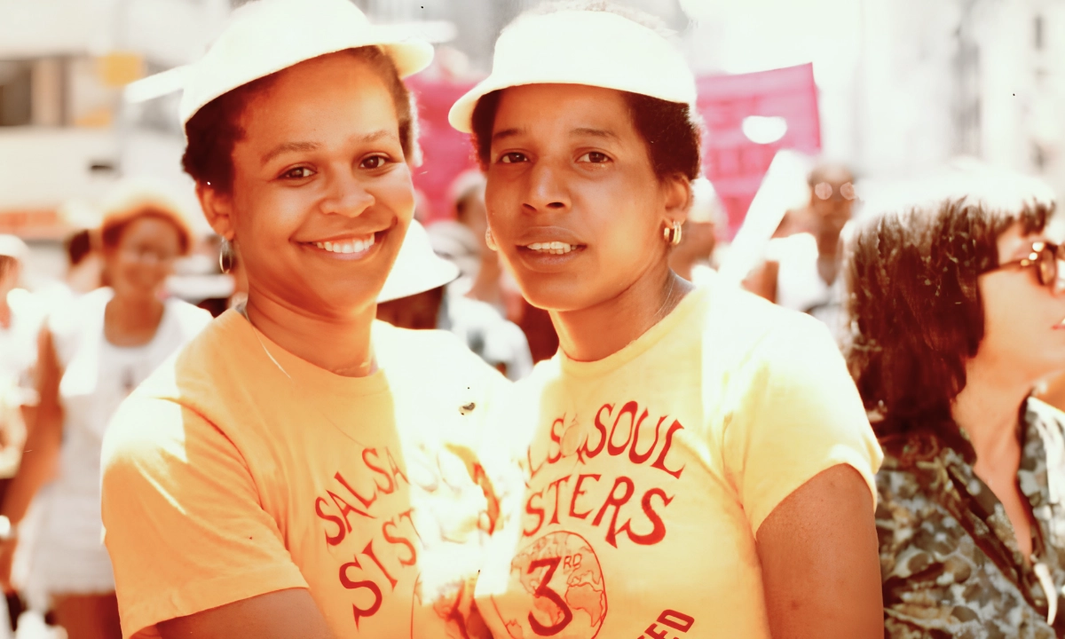 Two women wearing matching yellow t-shirts and hats stare into the camera and smile together in the middle of a crowd.