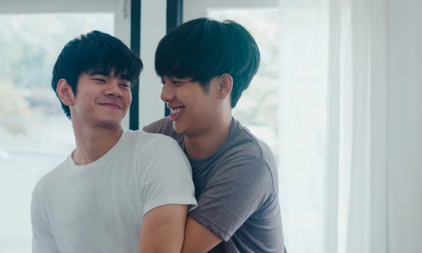 Gay man cuddles partner from behind as they both look and smile at each other.