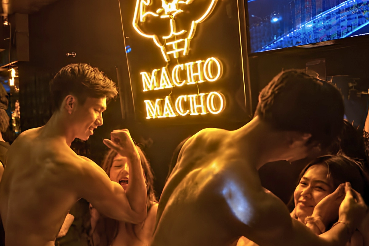 Two shirtless muscular performers interact with guests beneath a glowing “Macho Macho” sign.