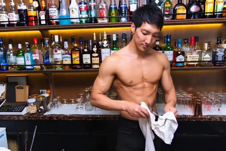Shirtless muscular bartender dries a glass behind a fully stocked bar.