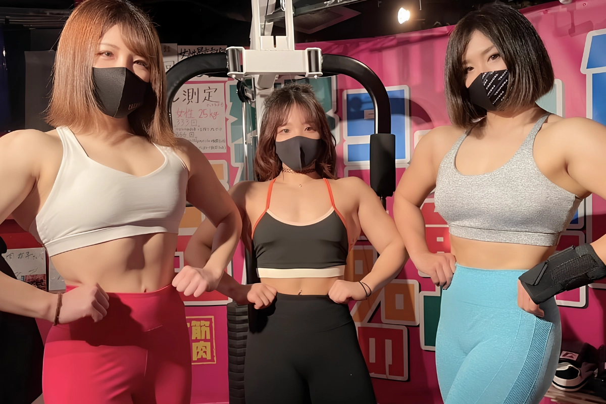 Three muscular women pose together in workout clothing inside a colorful muscle-themed venue.
