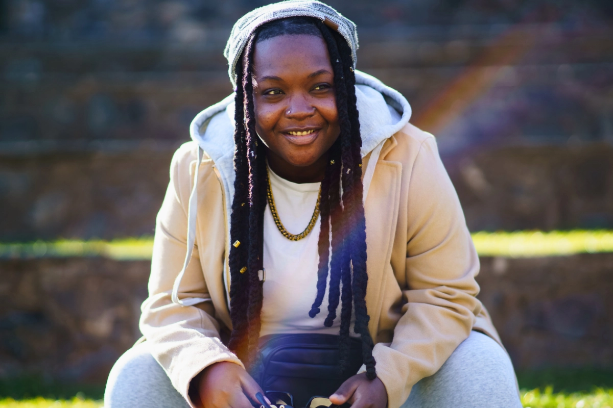 What Is a Stud Lesbian? The Black Queer History Behind the Identity 19 Smiling Black lesbian woman with braided dreadlocks wearing beanie looking away sitting with elbows leaning on knees outdoors on sunny day.