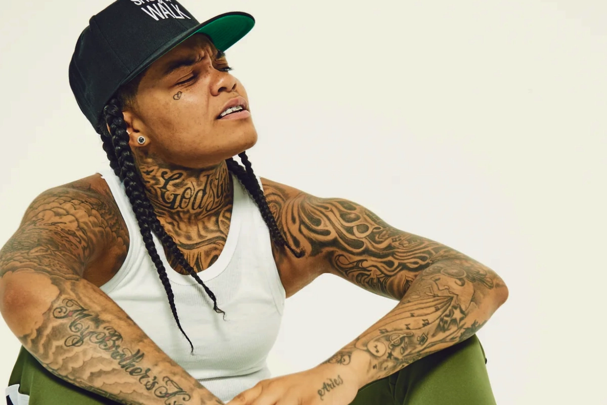 Tattooed person with long braids wears a black flat-brim cap, white tank top and green trousers while seated against a plain light background.
