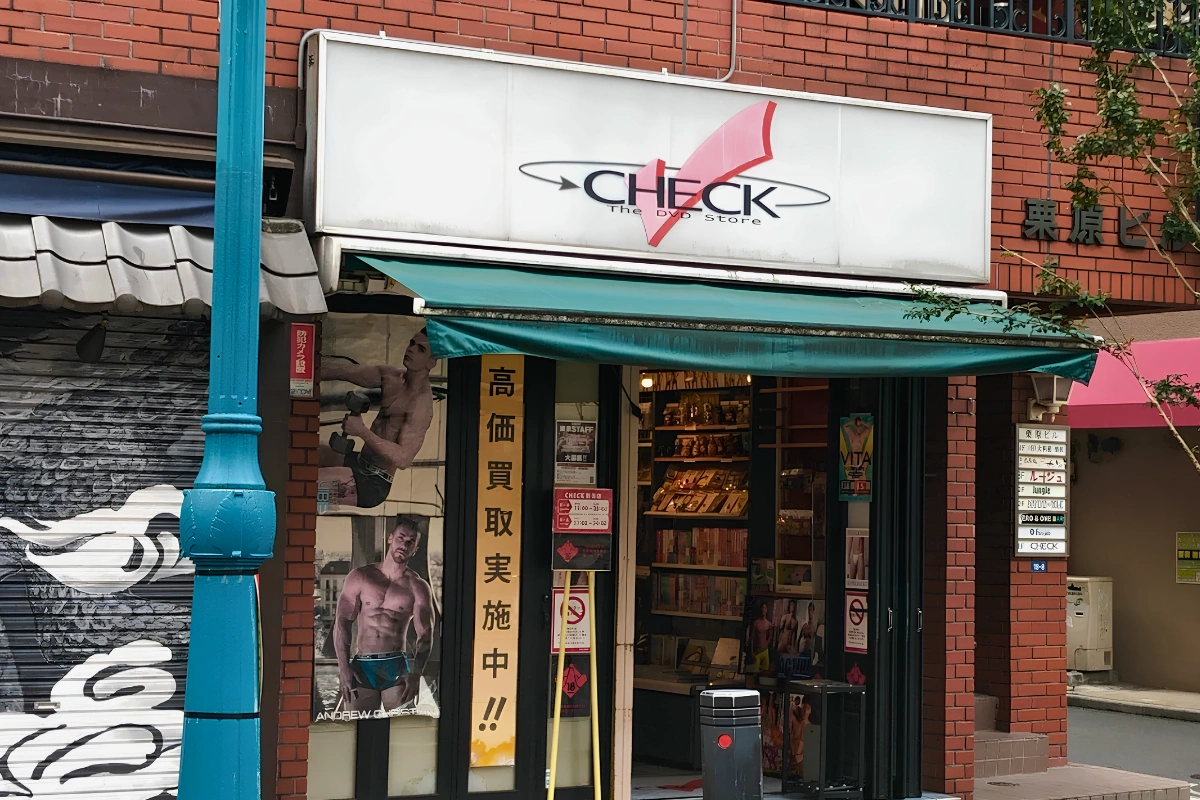 Exterior of CHECK, an adult store in Shinjuku Ni-chome, with its illuminated sign above the entrance and shelves of books and media visible inside.