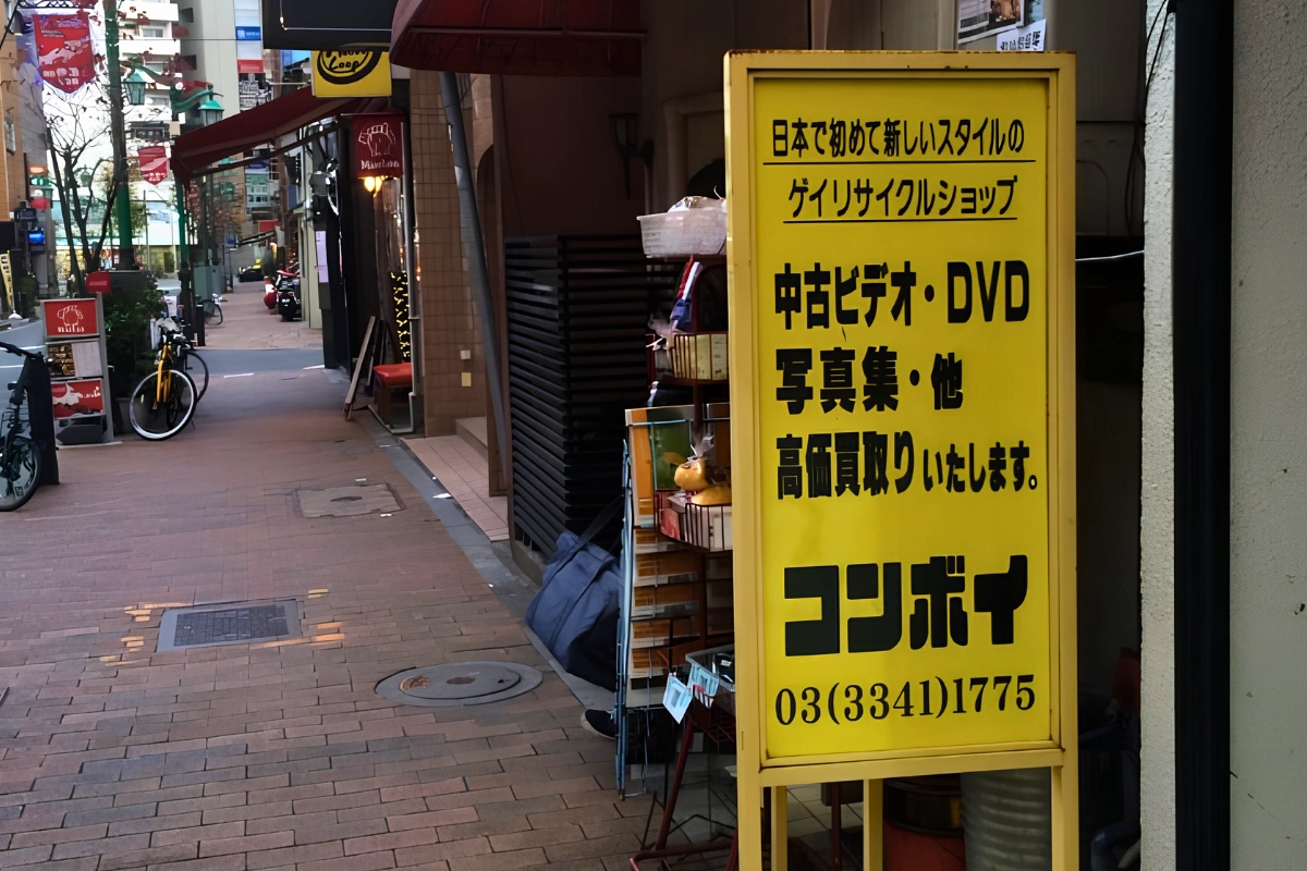 Yellow sidewalk sign outside a shop in Shinjuku Ni-chome advertising a gay resale store buying used videos, DVDs, and photo books, with the narrow shopping street visible behind it.