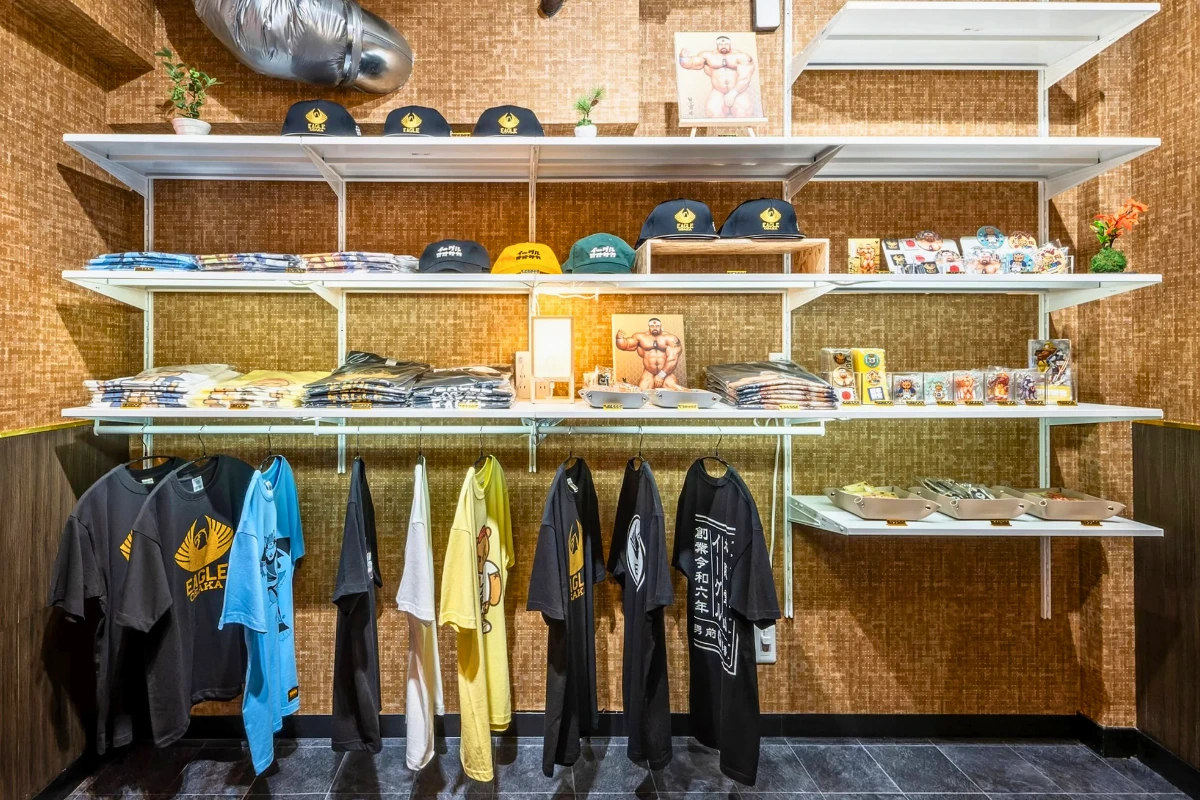 Merchandise display inside Eagle Osaka, with T-shirts, caps, pins, and small accessories arranged on shelves against a brown textured wall.