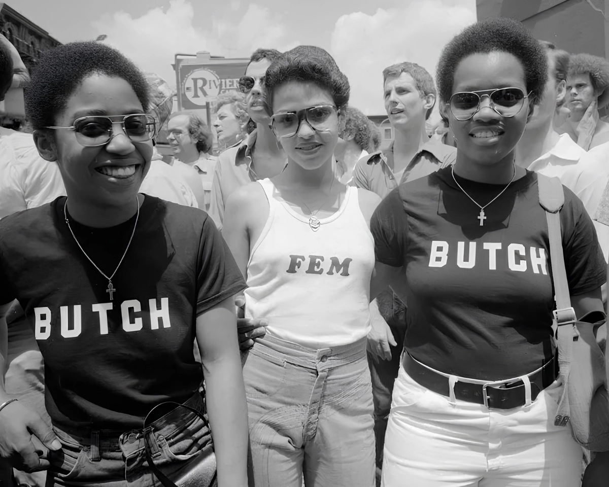 Black-and-white photograph of three people at a Pride march, with two wearing shirts reading “BUTCH” and the person in the center wearing a “FEM” shirt.