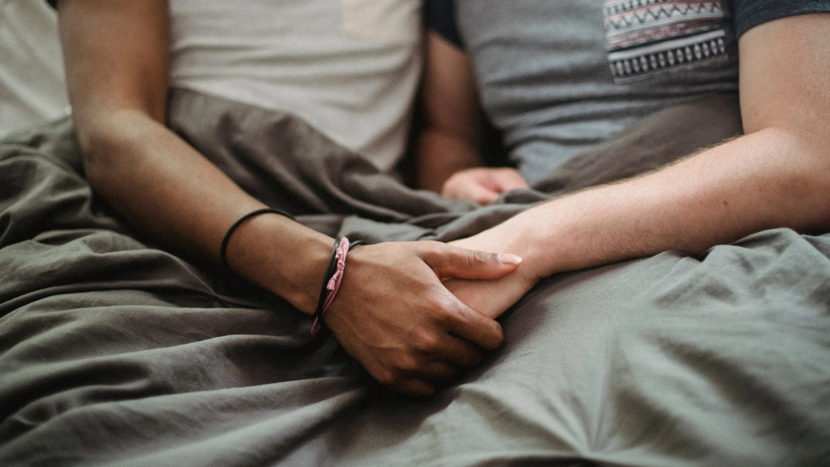 Two men holding hands intimately under blankets in bed, representing gay hookup culture and dating in Japan