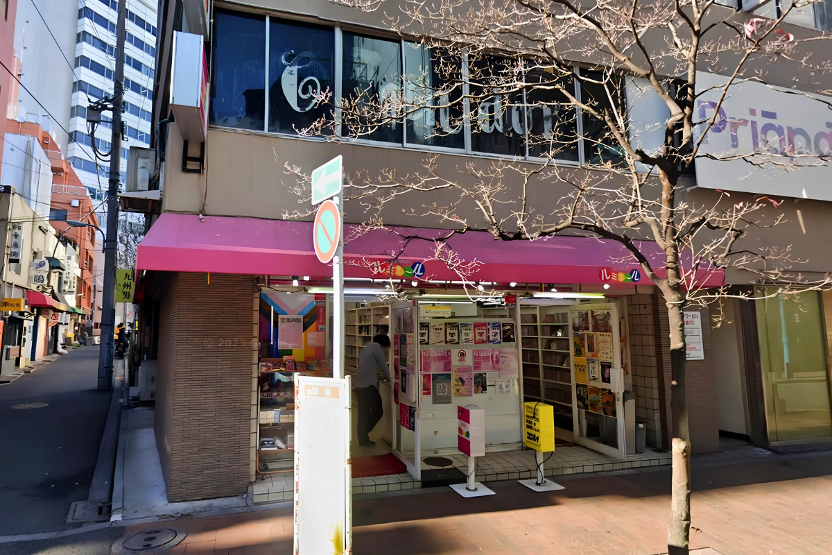 Daytime exterior of Lumiere in Shinjuku Ni-chome, with a bright pink awning, books and DVDs visible inside, and narrow side streets between surrounding buildings.