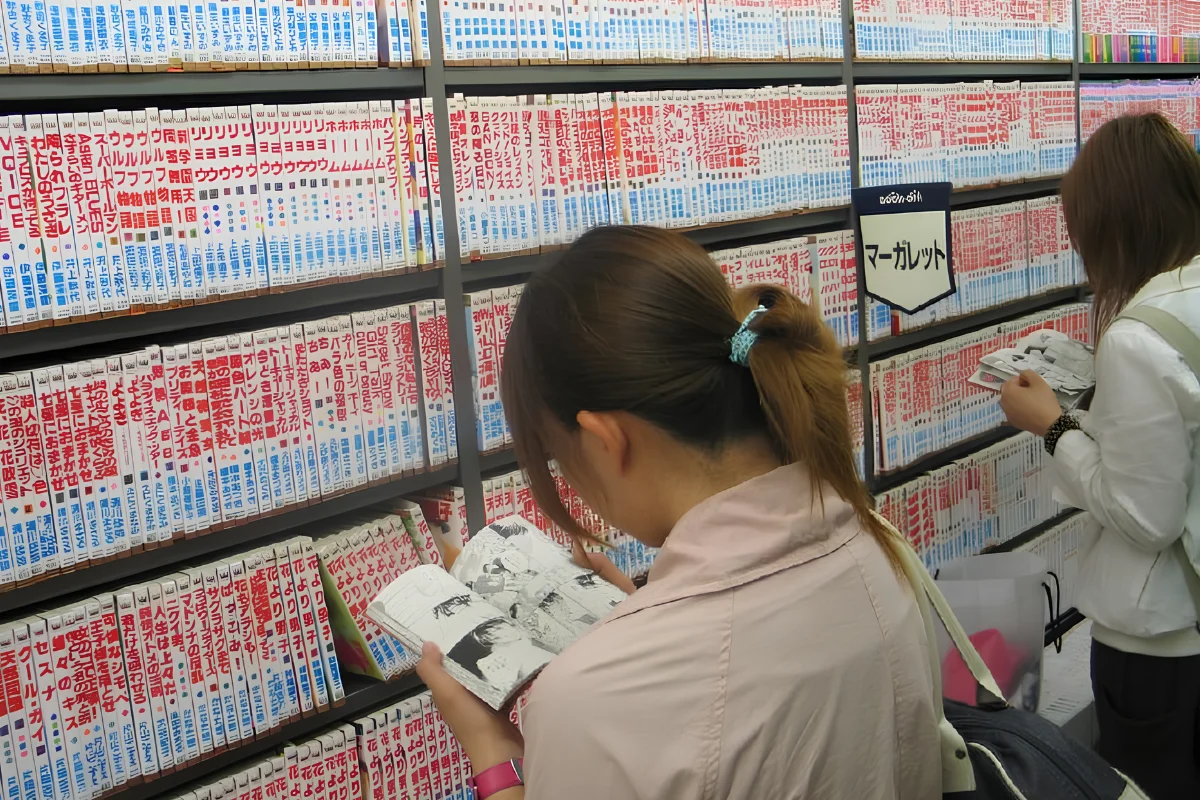 Two women in a bookstore in Japan read manga. The shelves are full of similar looking manga, all with a white spine and read writing showing the manga title.